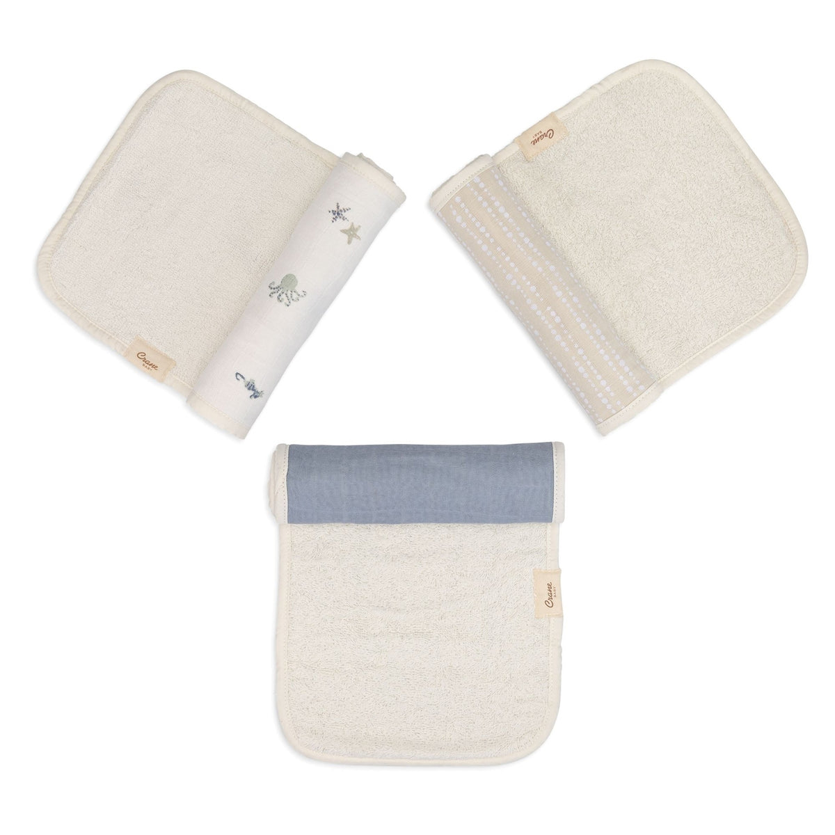 Three reusable baby wipes or cloths shown on a white background: one folded cream cloth with a rolled section showing small gray animal and star prints, one folded cream cloth with dotted trim, and one folded cream cloth with a dusty blue band near the top.