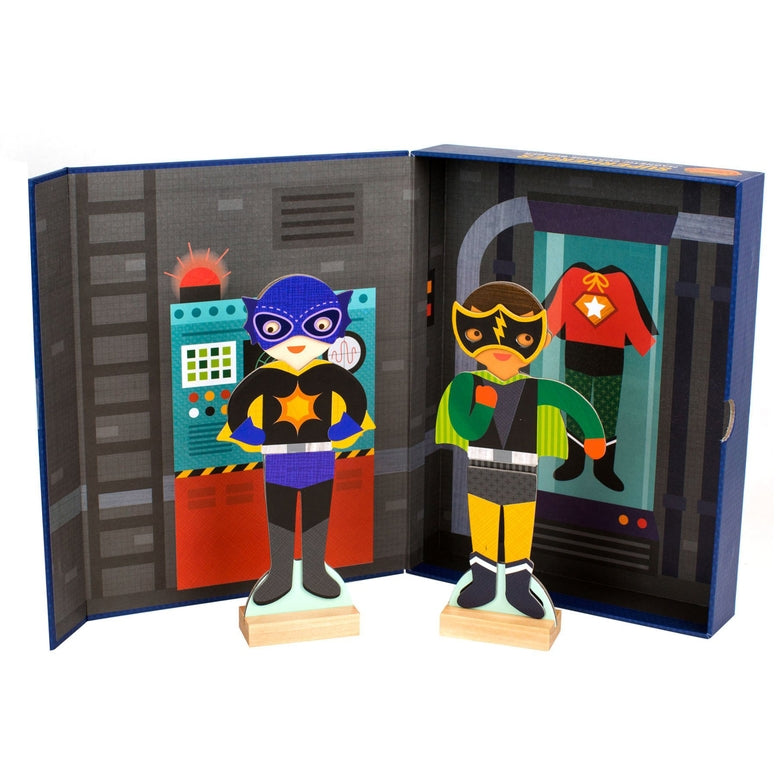 Two magnetic superhero figures from the Superheroes Magnetic Costume Builder by Petit Collage standing on wooden bases inside an open storage box decorated like a secret hideout.