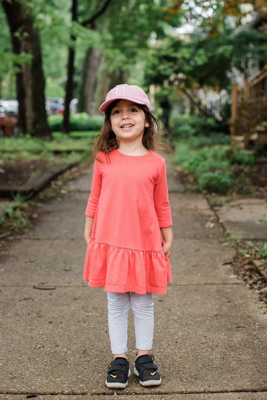Young girl in a pink dress and cap standing on a sidewalk with greenery in the background