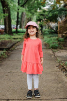 Young girl in a pink dress and cap standing on a sidewalk with greenery in the background