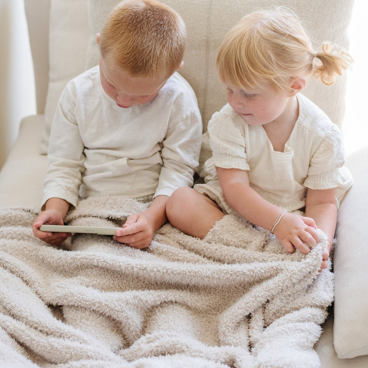 Two children sitting on a couch under a blanket, reading a book together.