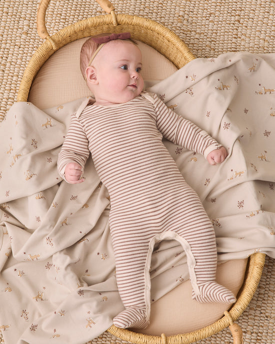 A baby lying on a cream and beige floral blanket inside a woven rattan bassinet, wearing a beige and brown striped footed sleeper.