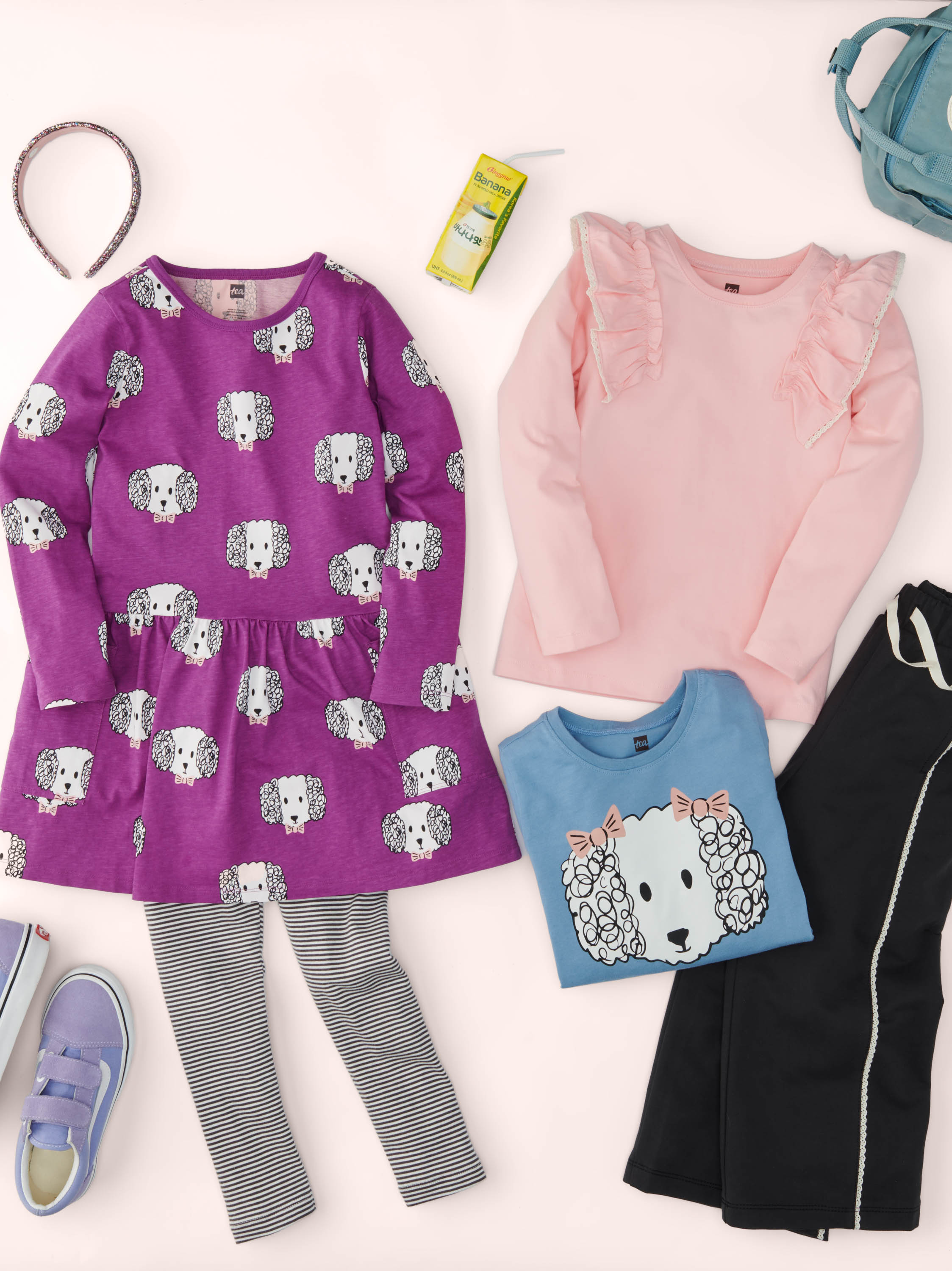 Children's clothing including a purple dress with dog pattern, pink sweatshirt, blue shirt with bear design, black pants, and shoes on a white background.