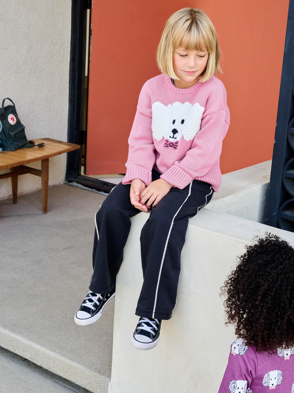 Child sitting on a couch wearing a pink sweater with a white bear design and black pants.