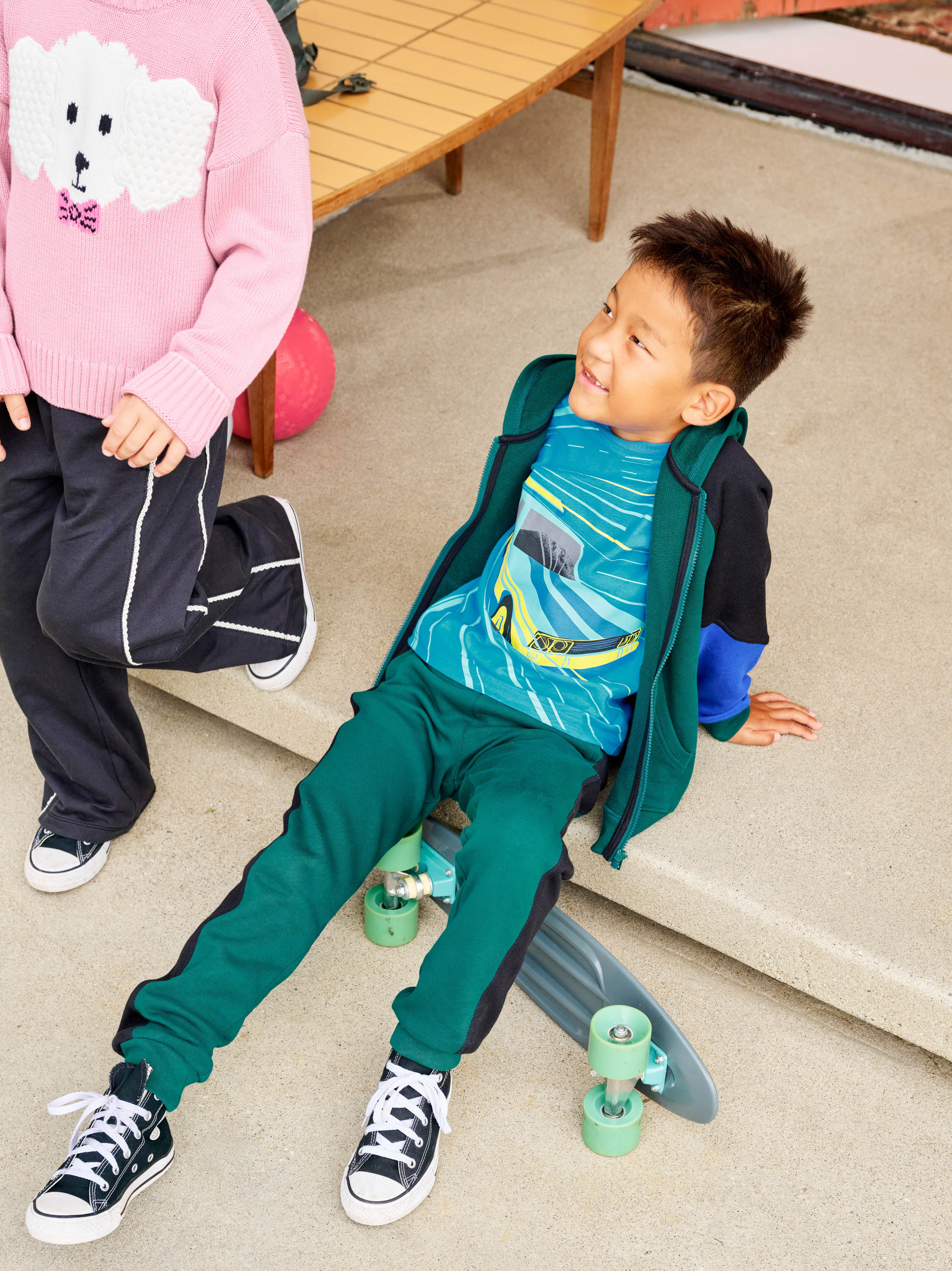 Child sitting on a skateboard with another child standing nearby, both indoors.