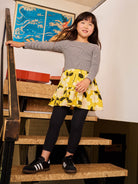Young girl in a yellow skirt and gray top standing on a staircase.