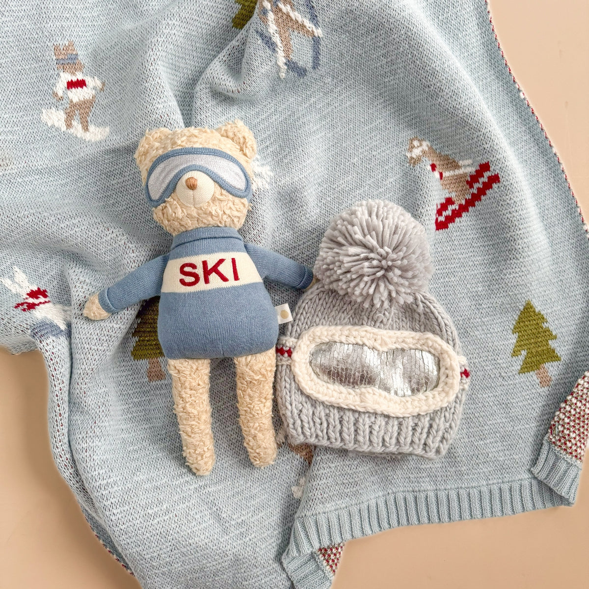 Teddy bear in ski outfit with goggles and a matching hat on a knitted blanket with winter patterns.