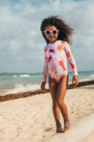 Child in a colorful swimsuit and sunglasses standing on a beach.