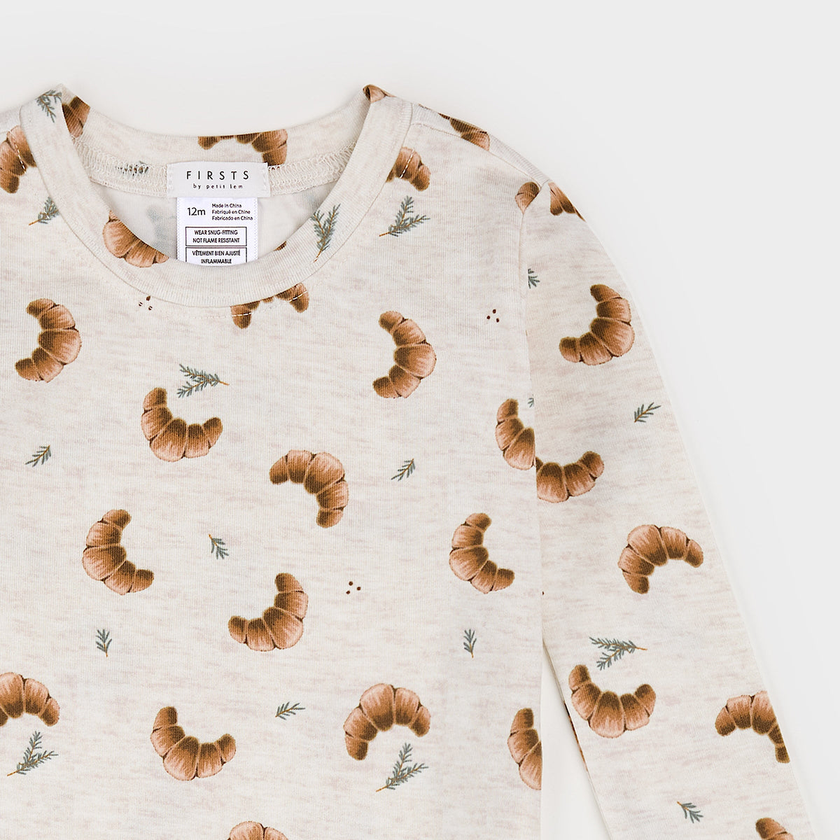 A close-up of a long-sleeve crewneck top by Firstst, featuring an all-over print of brown croissants and small green pine sprigs on a light cream background.