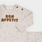 A baby long-sleeve top and matching pants set in a soft, light beige ribbed fabric, featuring a 'BON APPÉTIT' text design in brown sequins on the chest and a subtle repeating bow pattern. The brand 'Finsts' is visible on the inner neck label.