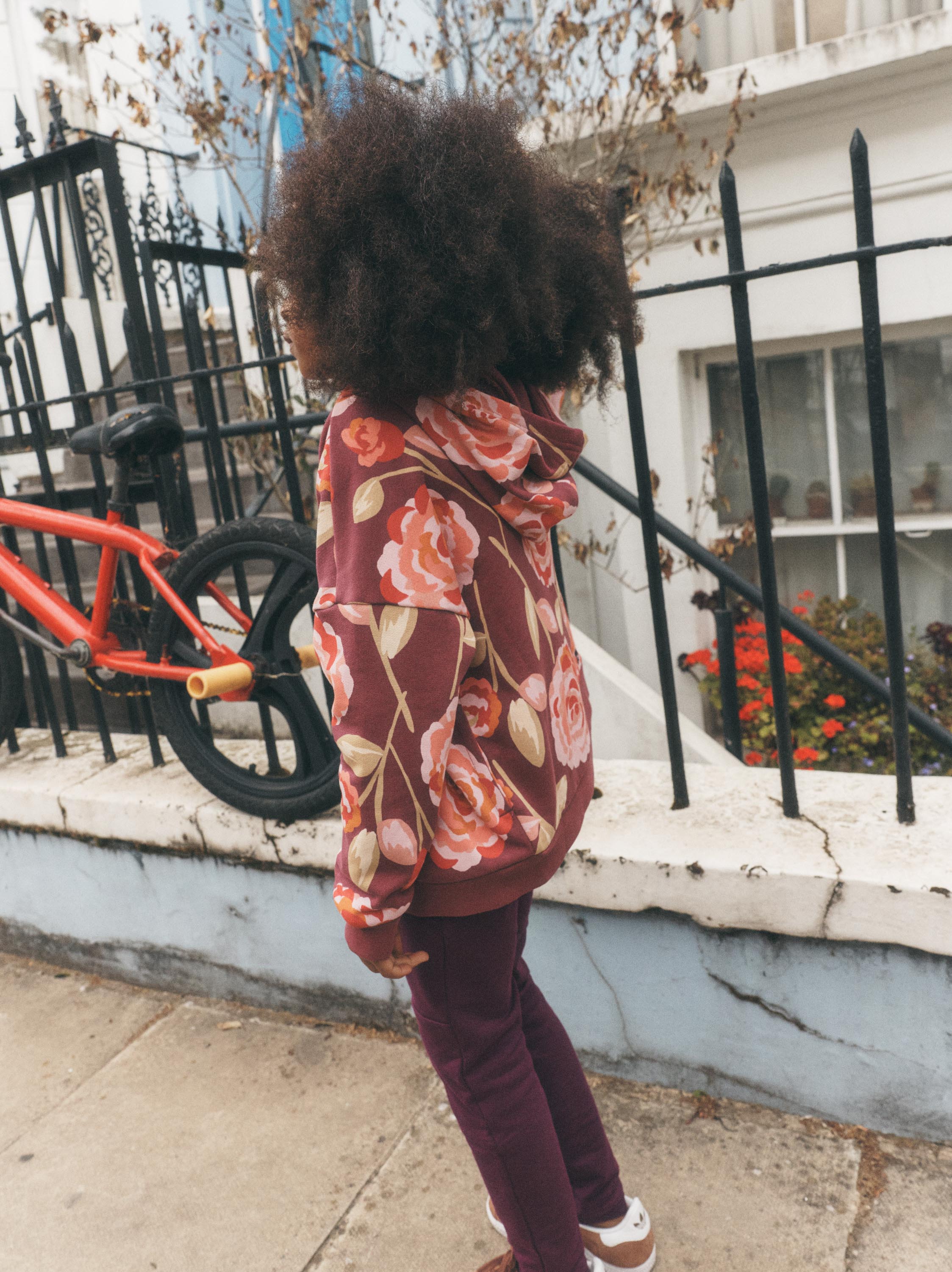 A child standing outdoors on a sidewalk in front of a white building and black metal fence, wearing a burgundy hoodie with a large pink and peach floral print and dark burgundy pants.