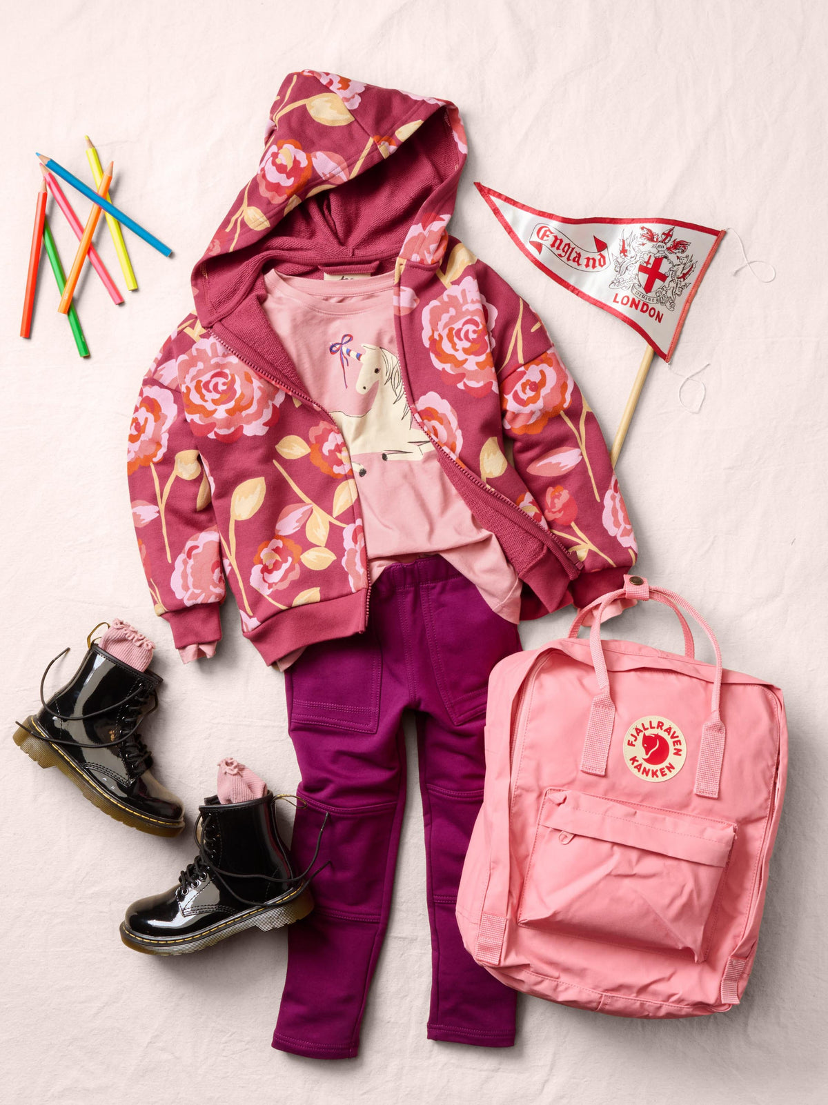 A styled flat-lay outfit featuring a pink floral hooded jacket, a light pink t-shirt, magenta pants, a pink Fjälör backpack, and black boots, arranged on a textured off-white background with pencils and a flag.