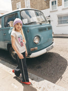 A young girl stands on a street curb in front of a vintage teal bus, wearing a light purple knit beanie, a white graphic t-shirt, dark wide-leg track pants with white side stripes, and bright red slip-on shoes.