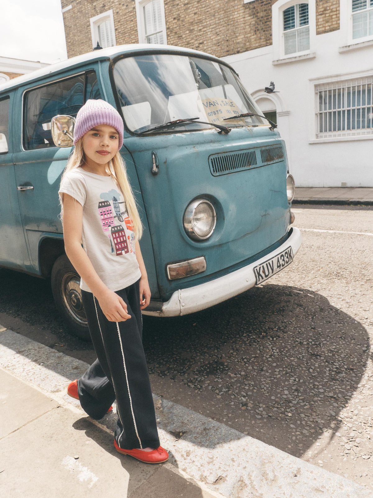 A young girl stands on a street curb in front of a vintage teal bus, wearing a light purple knit beanie, a white graphic t-shirt, dark wide-leg track pants with white side stripes, and bright red slip-on shoes.