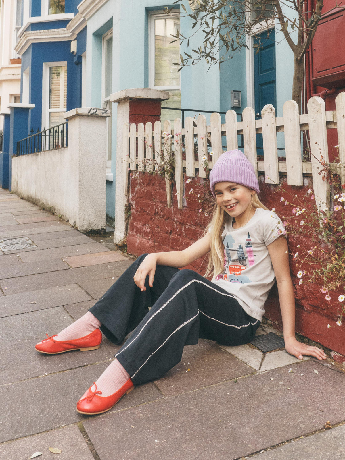 A young girl sitting on a sidewalk in front of a white picket fence and colorful houses. She is wearing a light purple knit beanie, a light gray graphic t-shirt, black wide-leg sweatpants with white side stripes, and bright red flat shoes.