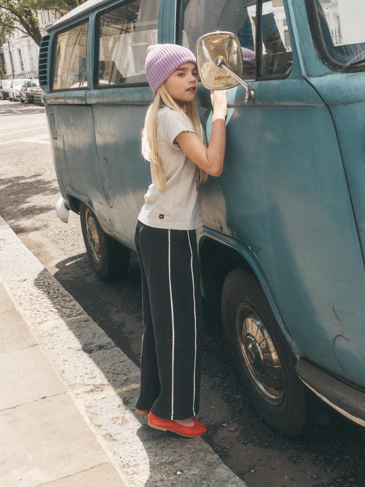 A young girl with long blonde hair stands next to a vintage teal van on a street corner, wearing a lavender knit beanie, a light gray t-shirt, black wide-leg track pants with white side stripes, and bright orange-red slip-on shoes.