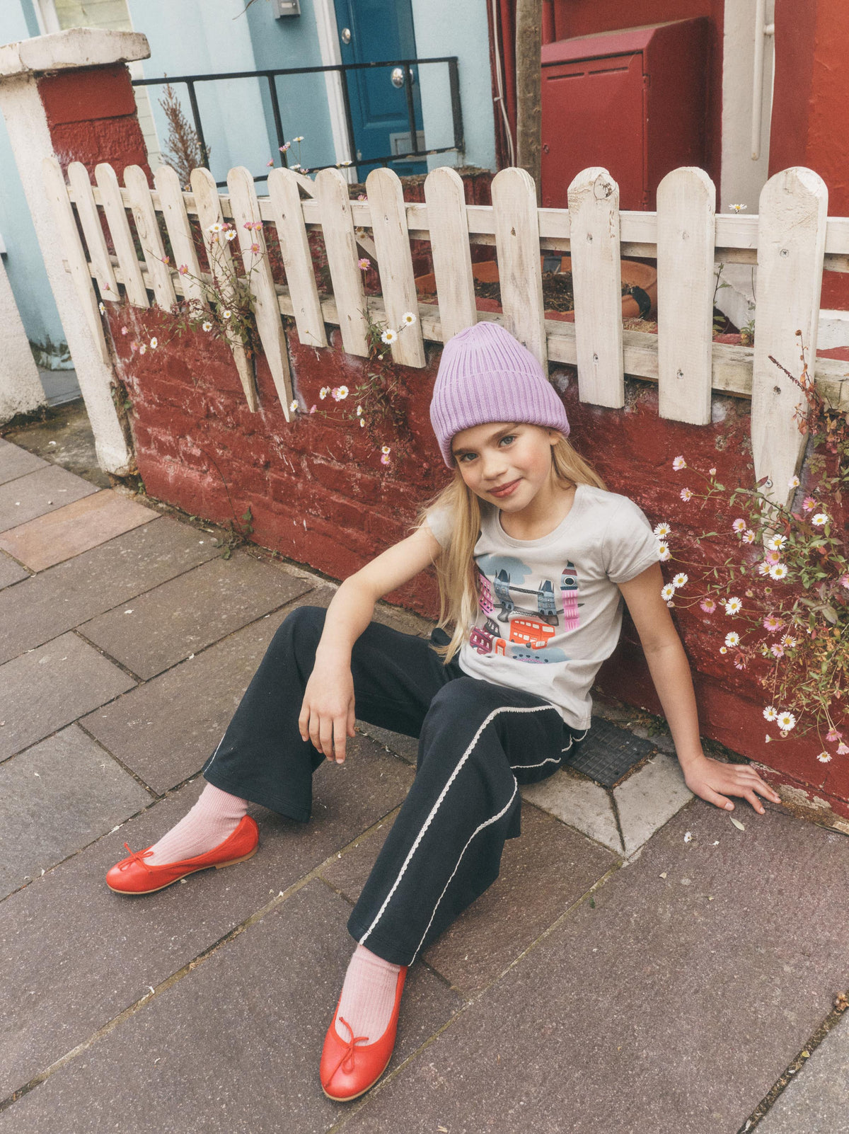 A young girl sitting on a brick sidewalk in front of a white picket fence and red brick wall, wearing a light purple knit beanie, a light gray graphic t-shirt, black track pants with white side stripes, and bright red shoes.