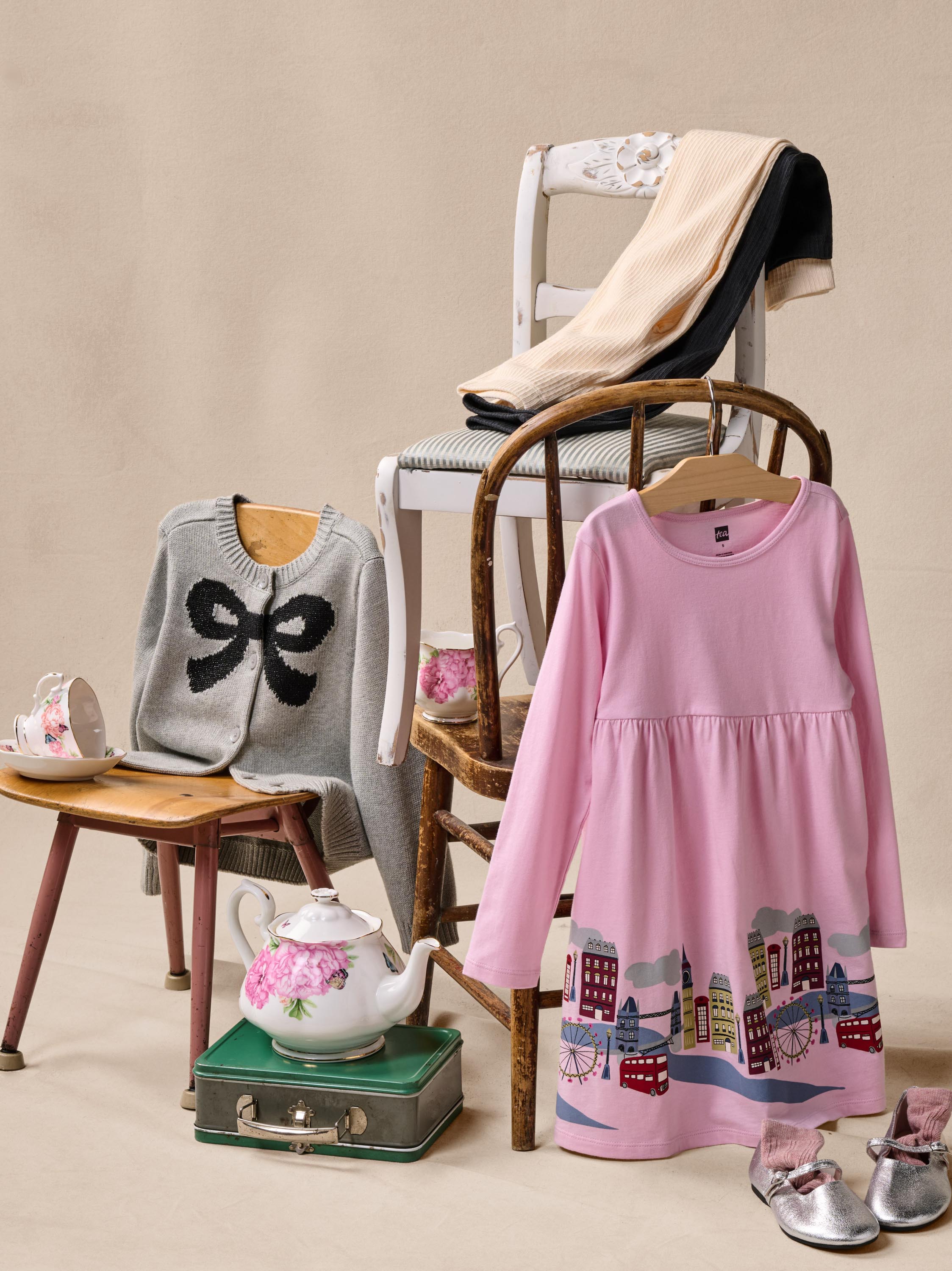 A styled product display featuring a pink long-sleeve babydoll dress with a cityscape print at the hem, alongside other children's clothing including a gray cardigan with a black bow, a beige and black knit layer, and silver shoes, arranged on chairs and a stool against a beige background.