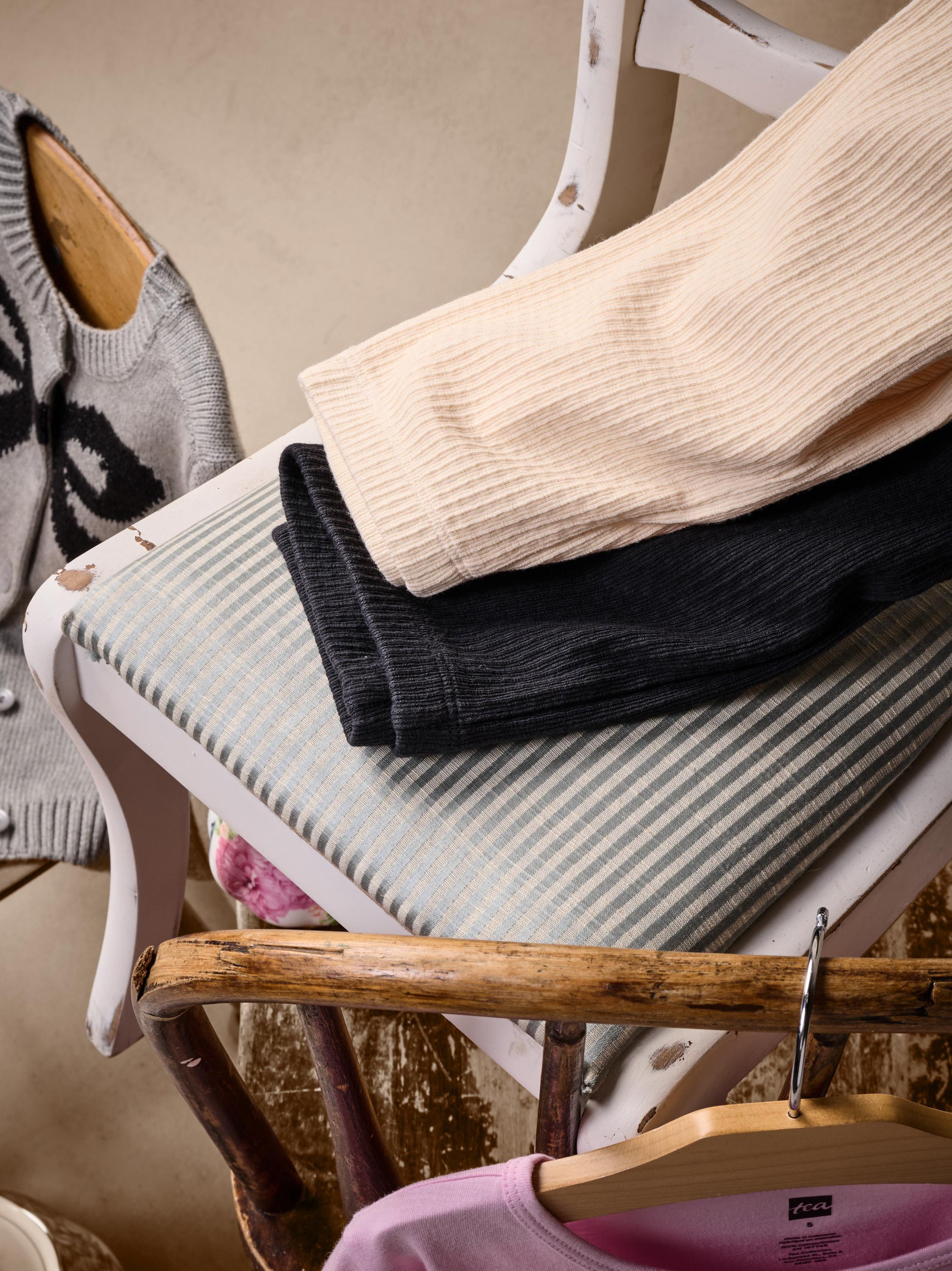 A styled product shot of folded ribbed knit garments on a white wooden chair. The stack includes a light beige ribbed top, a black ribbed bottom, and a gray-and-white striped cushion underneath.