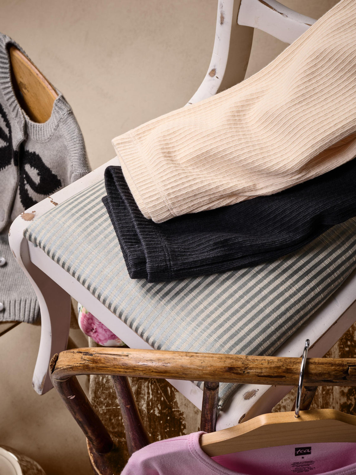 A styled product shot of folded ribbed knit garments on a white wooden chair. The stack includes a light beige ribbed top, a black ribbed bottom, and a gray-and-white striped cushion underneath.