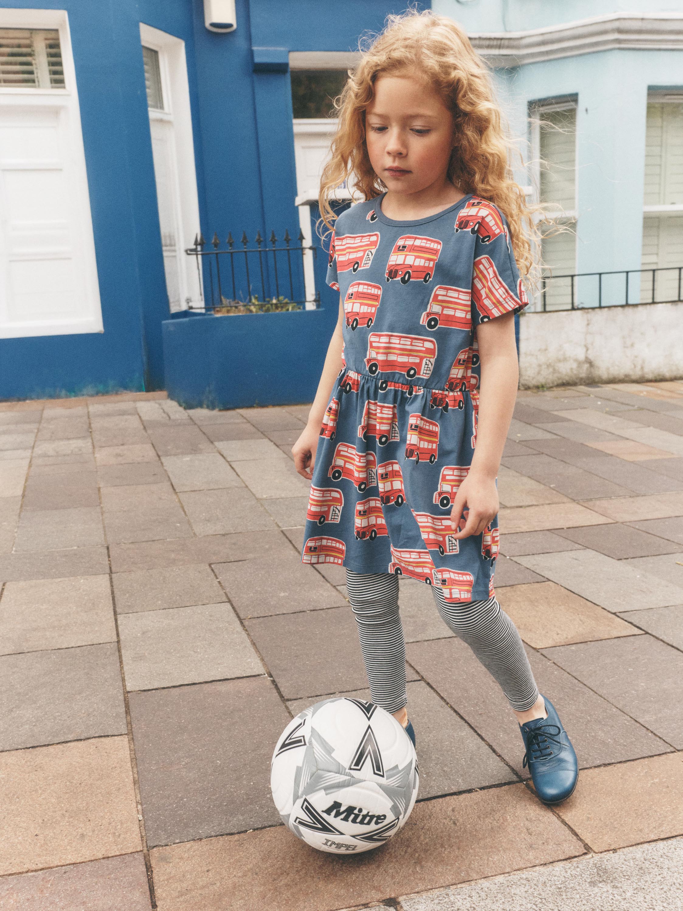 A young girl with curly blonde hair stands on a paved street in front of a blue house, wearing a blue dress with a red double-decker bus pattern, grey leggings, and blue shoes while balancing a soccer ball.