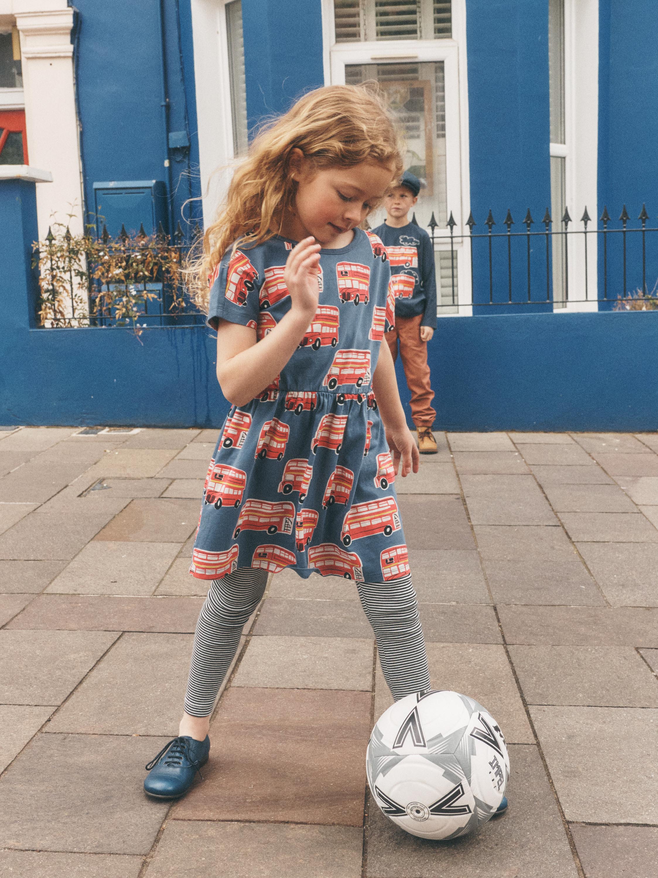 A young girl wearing a blue short-sleeve dress with a red and white bus print, grey leggings, and blue shoes is playing with a soccer ball on a paved street in front of a blue building.