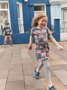 A young girl with curly hair smiles while walking outdoors on a paved street in front of a blue house. She is wearing a blue short-sleeve dress covered in red double-decker bus prints, gray striped leggings, and blue shoes.
