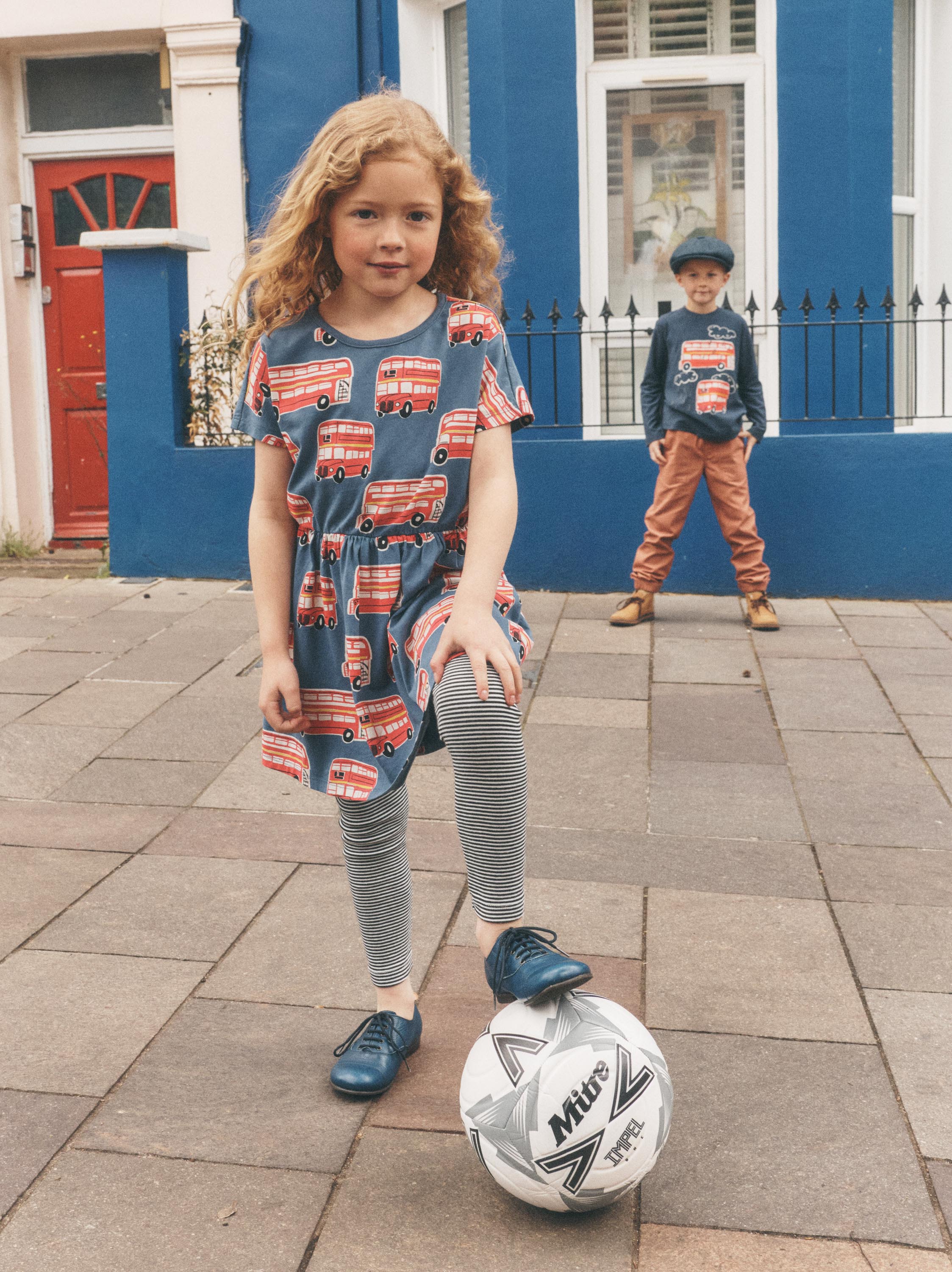 A young girl stands on a paved street in front of a blue building, wearing a blue short-sleeve dress with a red double-decker bus pattern, gray striped leggings, and blue shoes while balancing a soccer ball. A boy stands further back in casual outdoor clothing.