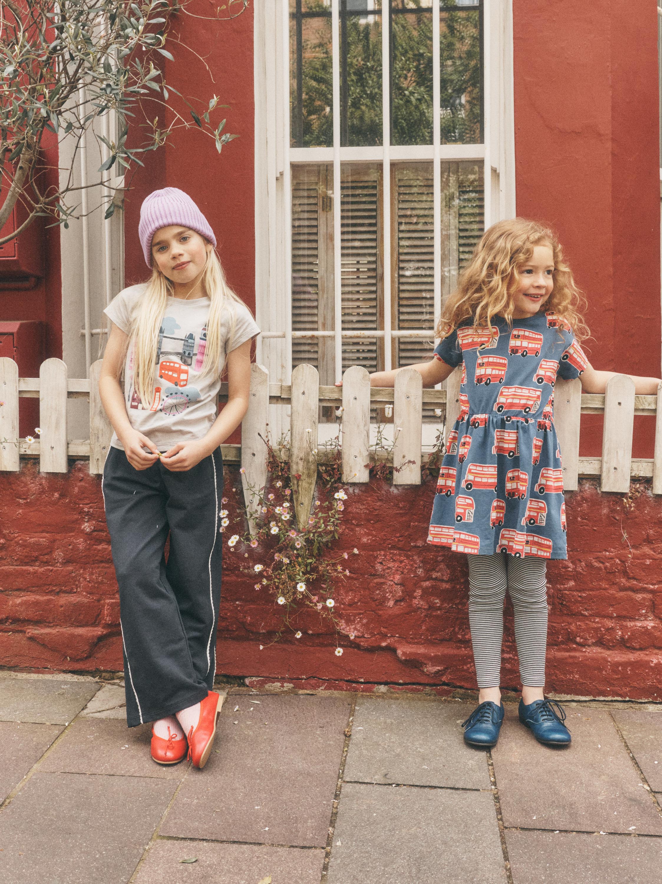 Two young girls standing outdoors in front of a red brick wall and white window, wearing casual clothing. The girl on the left wears a light purple beanie, a light graphic T-shirt, dark wide-leg pants, and red shoes.