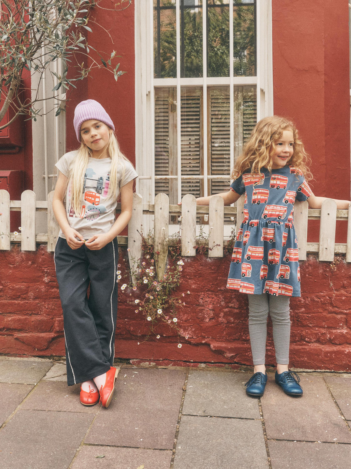 Two young girls standing outdoors in front of a red brick wall and white window, wearing casual clothing. The girl on the left wears a light purple beanie, a light graphic T-shirt, dark wide-leg pants, and red shoes.