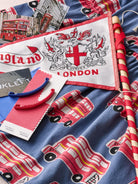 A close-up of a blue fabric product printed with red London buses and a white pennant flag featuring a red cross, dragon, and the words 'England LONDON'. A red double-decker bus illustration and a red-and-white striped stick are also visible.