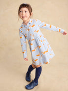 A young girl modeling a light blue long-sleeve dress with an orange and white fox print, paired with navy blue knee-high socks and navy shoes, against a warm beige studio backdrop.