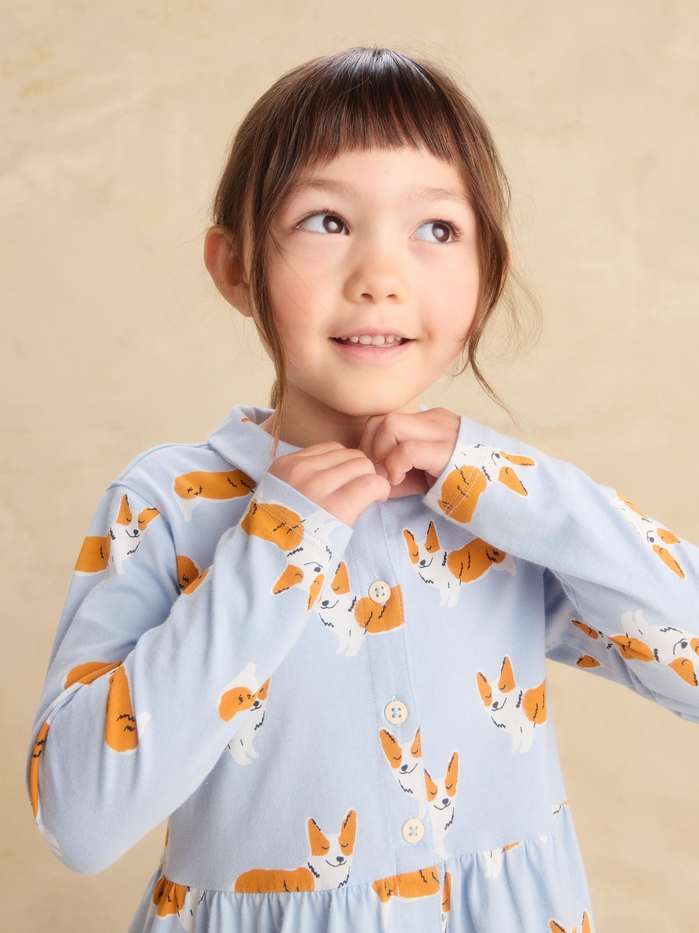 A young girl with brown hair and bangs wearing a light blue long-sleeve button-up shirt with an orange and white corgi dog print, posing against a warm beige background.