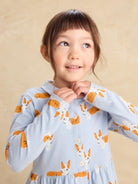 A young girl with brown hair and bangs wearing a light blue long-sleeve button-up shirt with an orange and white corgi dog print, posing against a warm beige background.