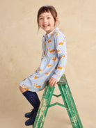 A young girl with brown hair sits on a distressed green wooden step ladder, wearing a light blue long-sleeve dress with an orange bunny print, dark blue knee-high socks, and navy boots against a textured beige studio backdrop.