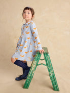 A young girl wearing a light blue long-sleeve dress with a yellow and white rabbit print, navy blue knee-high socks, and navy boots, posing on a green wooden step ladder against a textured beige studio backdrop.