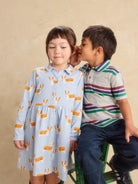 A young girl in a light blue collared dress with a corgi print sits next to a boy in a striped polo shirt who is whispering in her ear, set against a warm beige background.