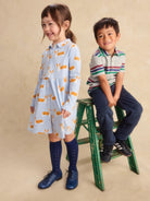 A young girl stands wearing a light blue long-sleeve dress with a yellow and white animal print, paired with dark blue knee-high socks and blue shoes. A young boy sits on a green wooden step stool wearing a striped polo shirt, navy pants, and black sneakers.