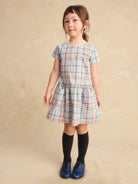 A young girl modeling a short-sleeved plaid dress with a tiered skirt, paired with black knee-high socks and blue shoes, standing against a textured beige studio backdrop.