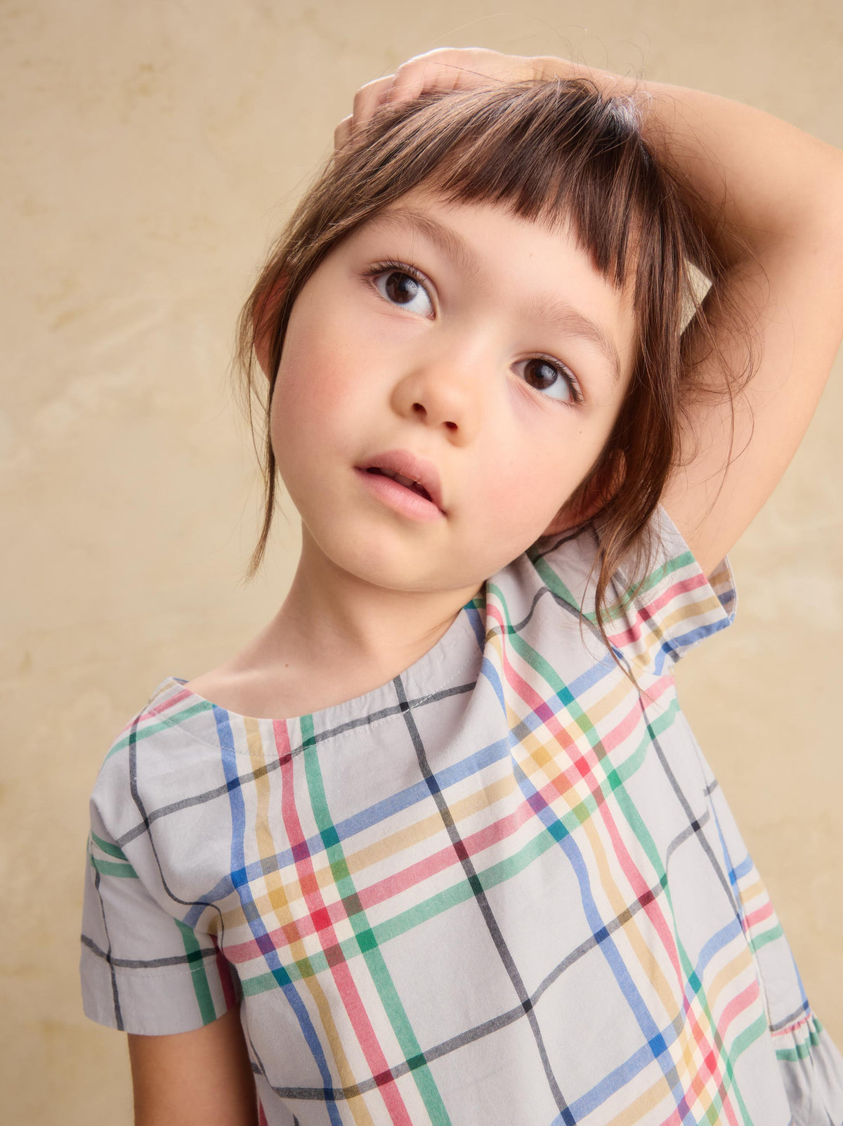A young girl with brown hair and bangs looks up and to the side while touching her hair, wearing a short-sleeve plaid top with a gray base and multicolor lines on a beige background.