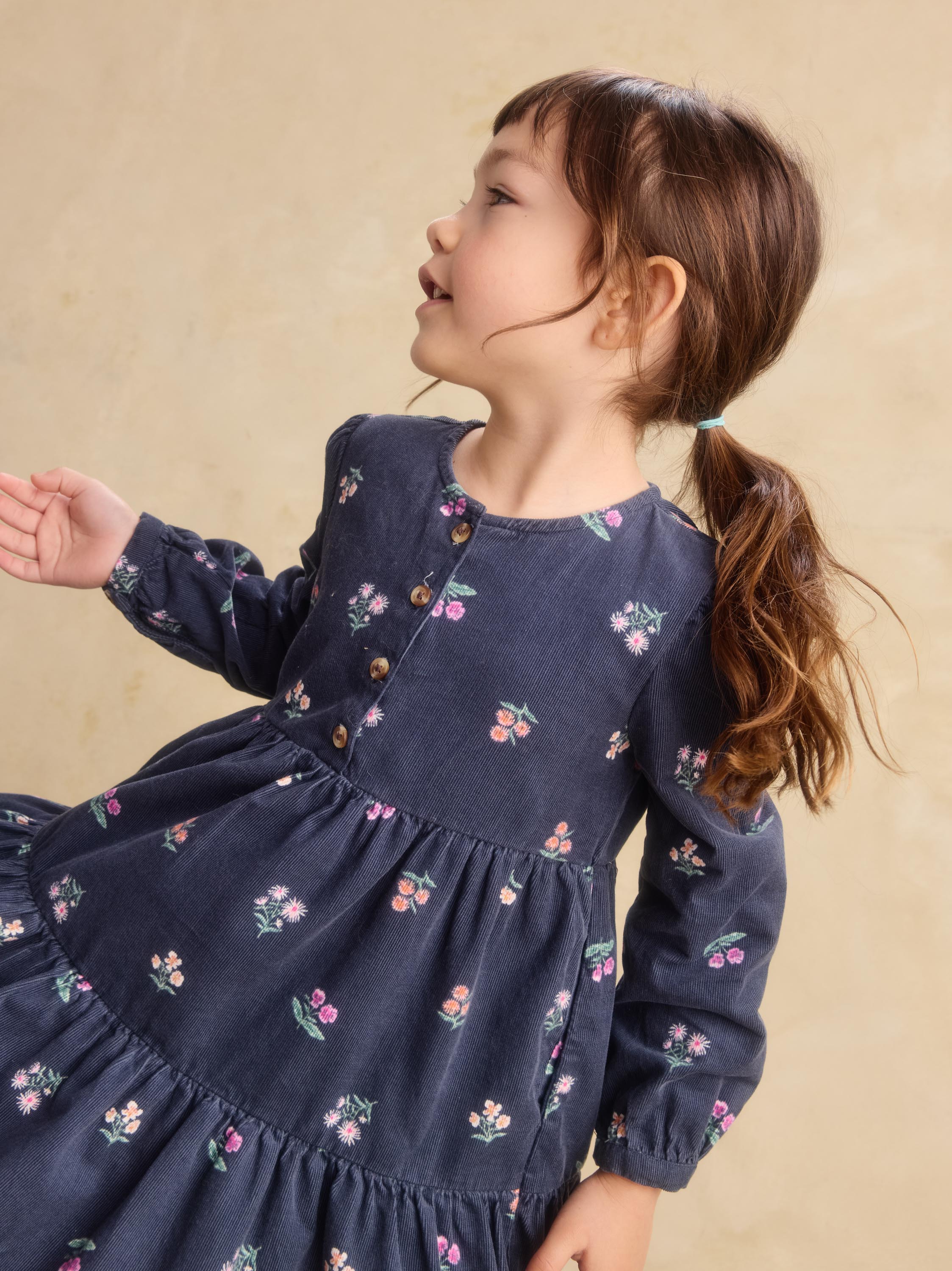 A young girl with brown hair in a ponytail wearing a navy blue long-sleeve tiered dress with a small pink and green floral print, posed against a warm beige background.