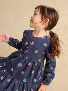 A young girl with brown hair in a ponytail wearing a navy blue long-sleeve tiered dress with a small pink and green floral print, posed against a warm beige background.