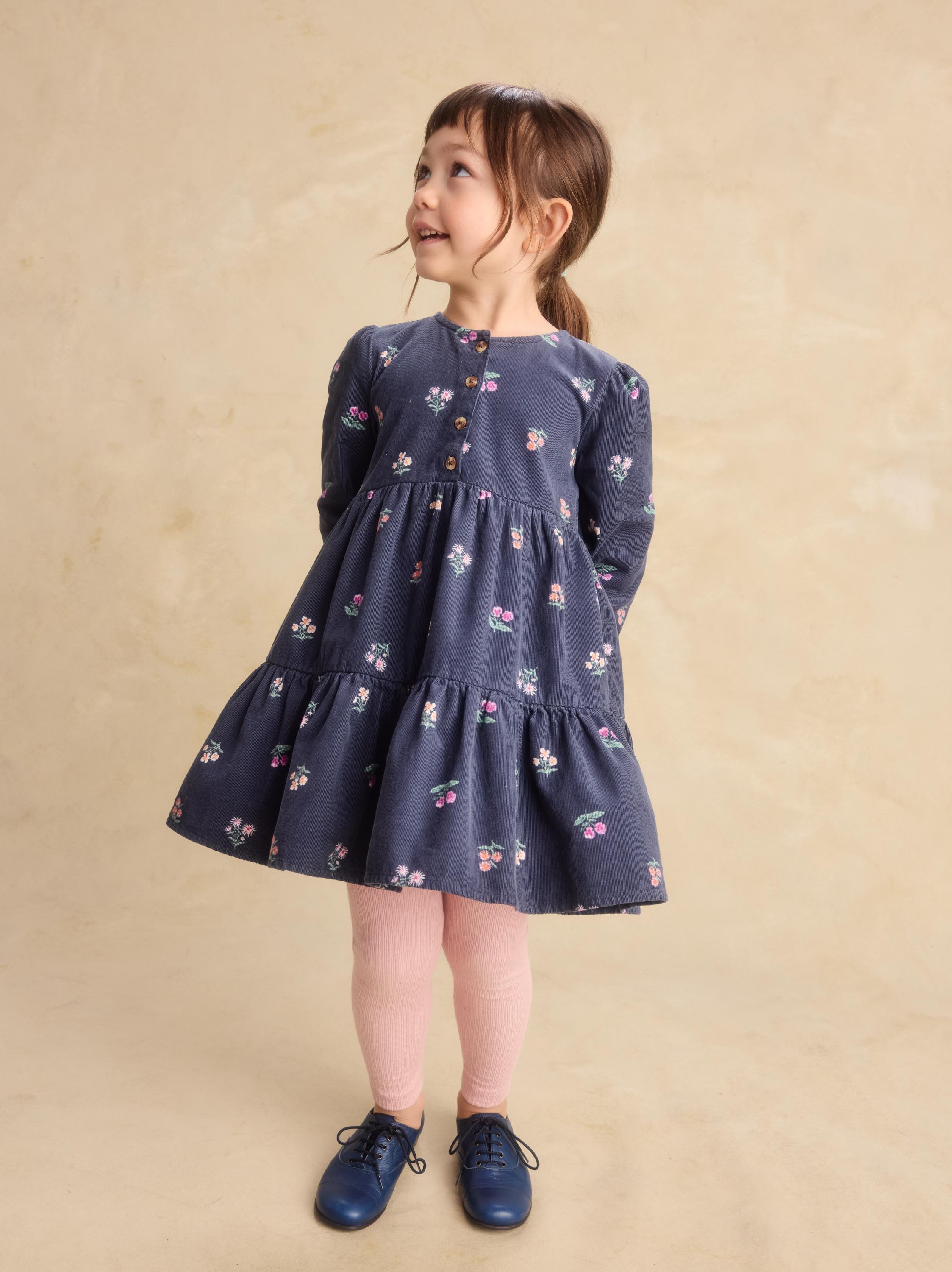 A young girl with brown hair in a ponytail stands wearing a navy blue tiered dress with a small floral print, pink tights, and navy shoes against a textured beige background.