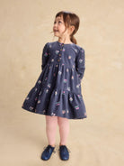 A young girl with brown hair in a ponytail stands wearing a navy blue tiered dress with a small floral print, pink tights, and navy shoes against a textured beige background.