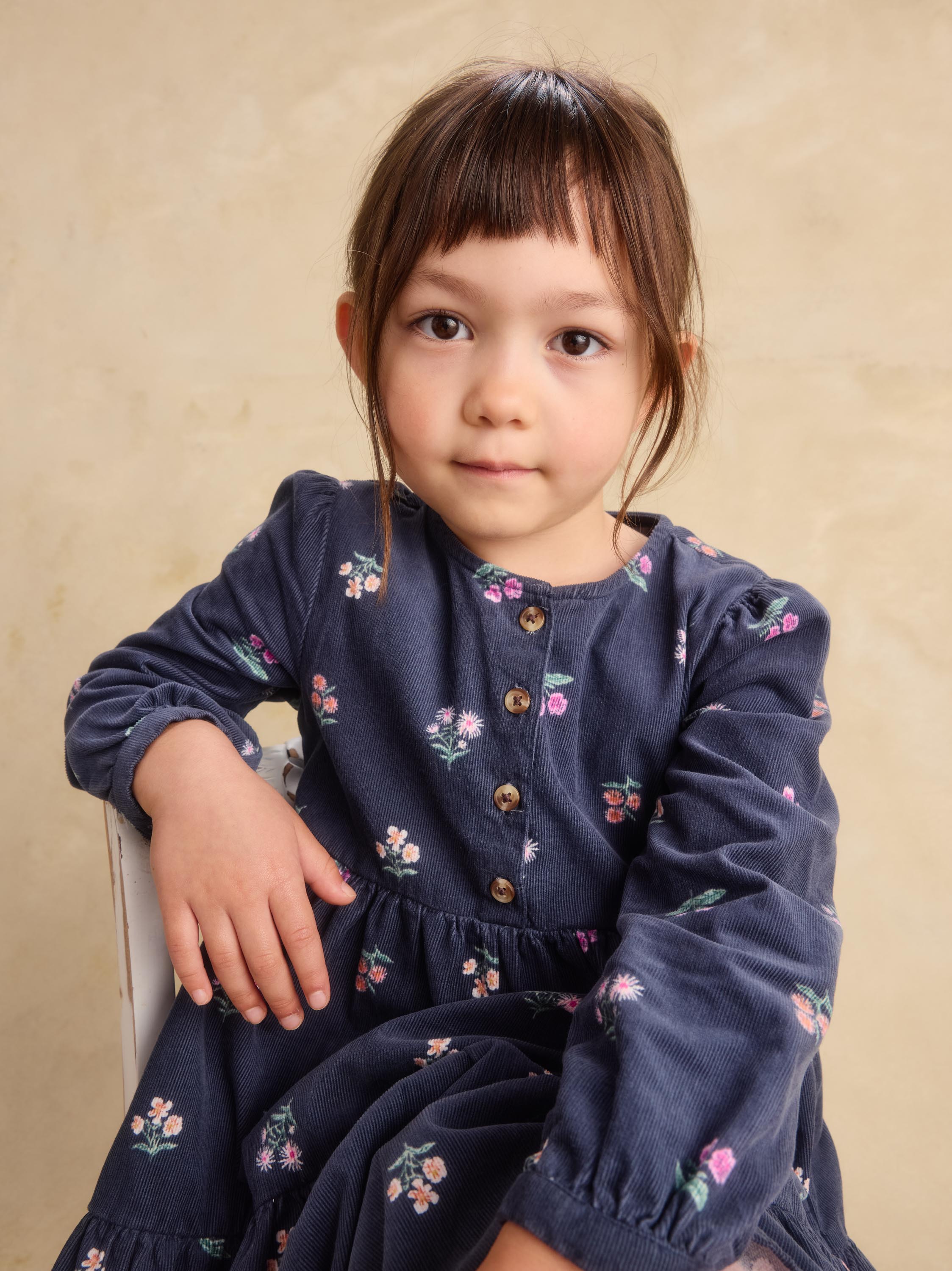 A young girl with dark brown hair and bangs wearing a navy blue corduroy dress with a small pink and green floral print, sitting against a textured beige background.