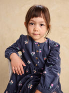 A young girl with dark brown hair and bangs wearing a navy blue corduroy dress with a small pink and green floral print, sitting against a textured beige background.