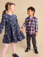 A young girl in a navy blue floral long-sleeve dress and a boy in a red, blue, and white plaid shirt are holding hands against a warm beige studio backdrop.