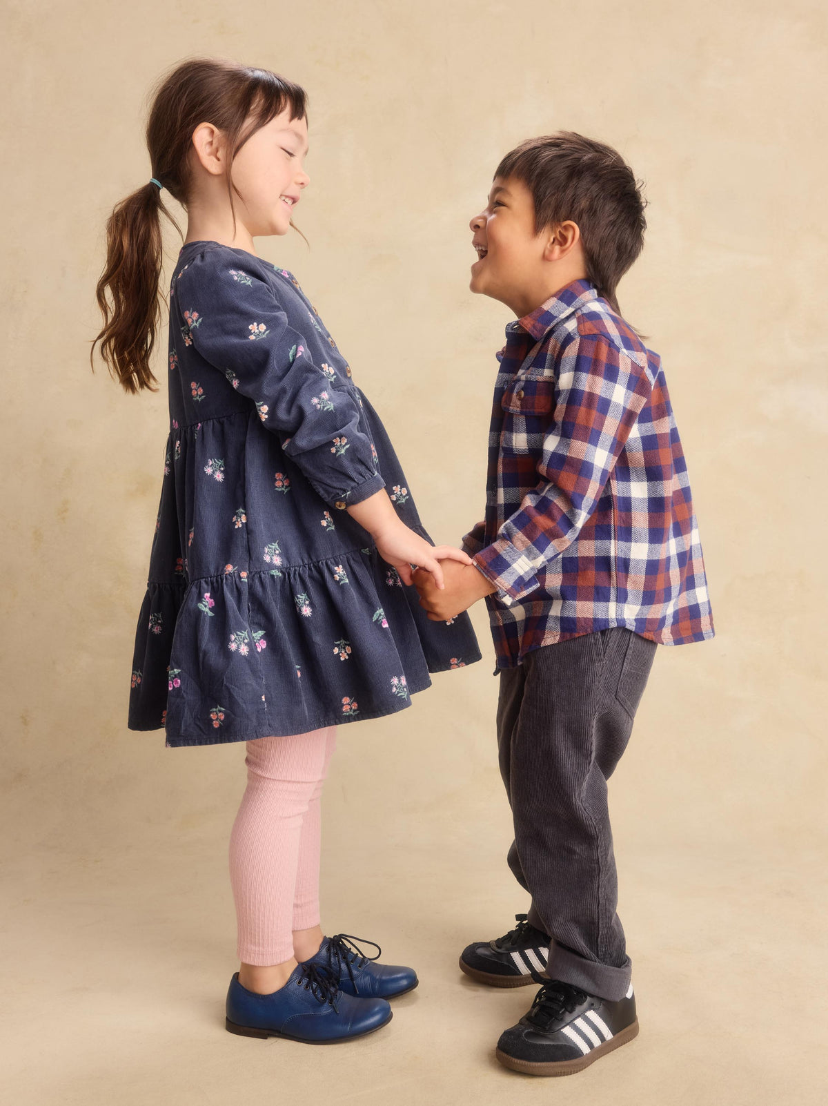 A young girl in a navy blue floral dress and pink leggings holds hands with a young boy wearing a red, blue, and white plaid shirt, dark gray pants, and black sneakers with white stripes, set against a warm beige studio backdrop.