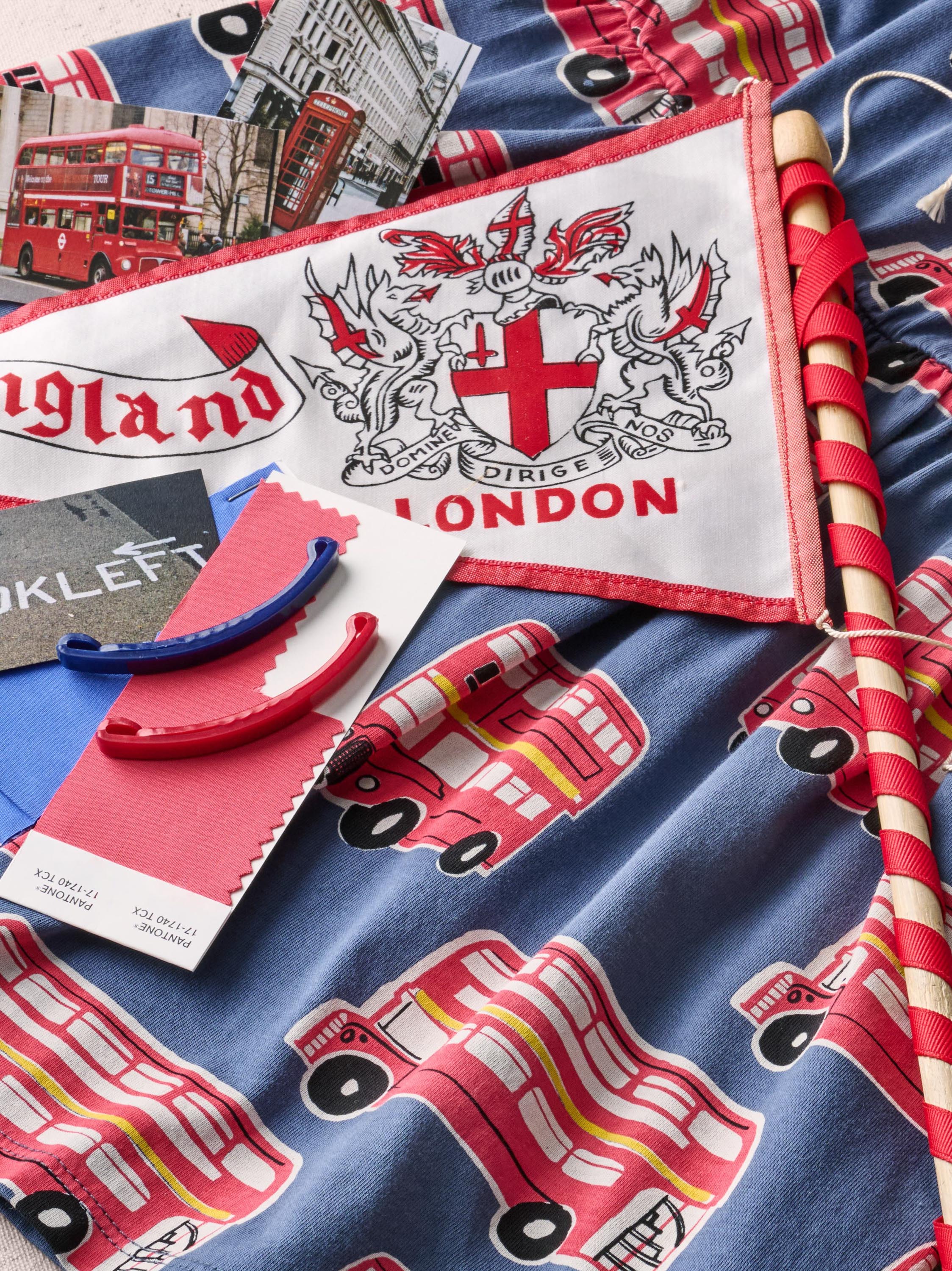 A close-up of a blue fabric product printed with red London buses and a white pennant flag featuring a red cross, dragon, and the words 'England LONDON'. A red double-decker bus illustration and a red-and-white striped stick are also visible.