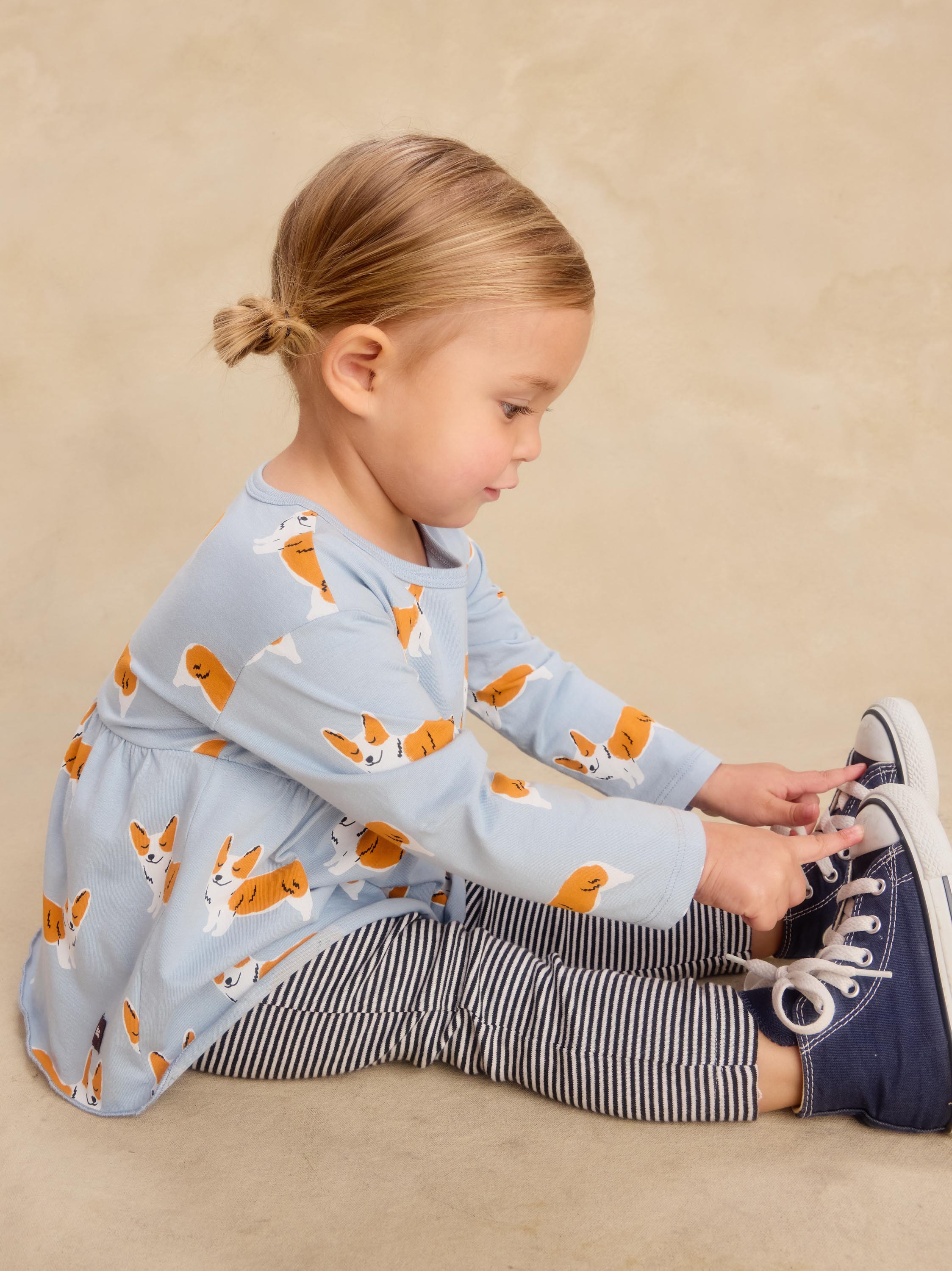 A toddler girl sitting on a beige studio floor, wearing a light blue long-sleeve top with an orange and white dog print, navy and white striped leggings, and navy sneakers.